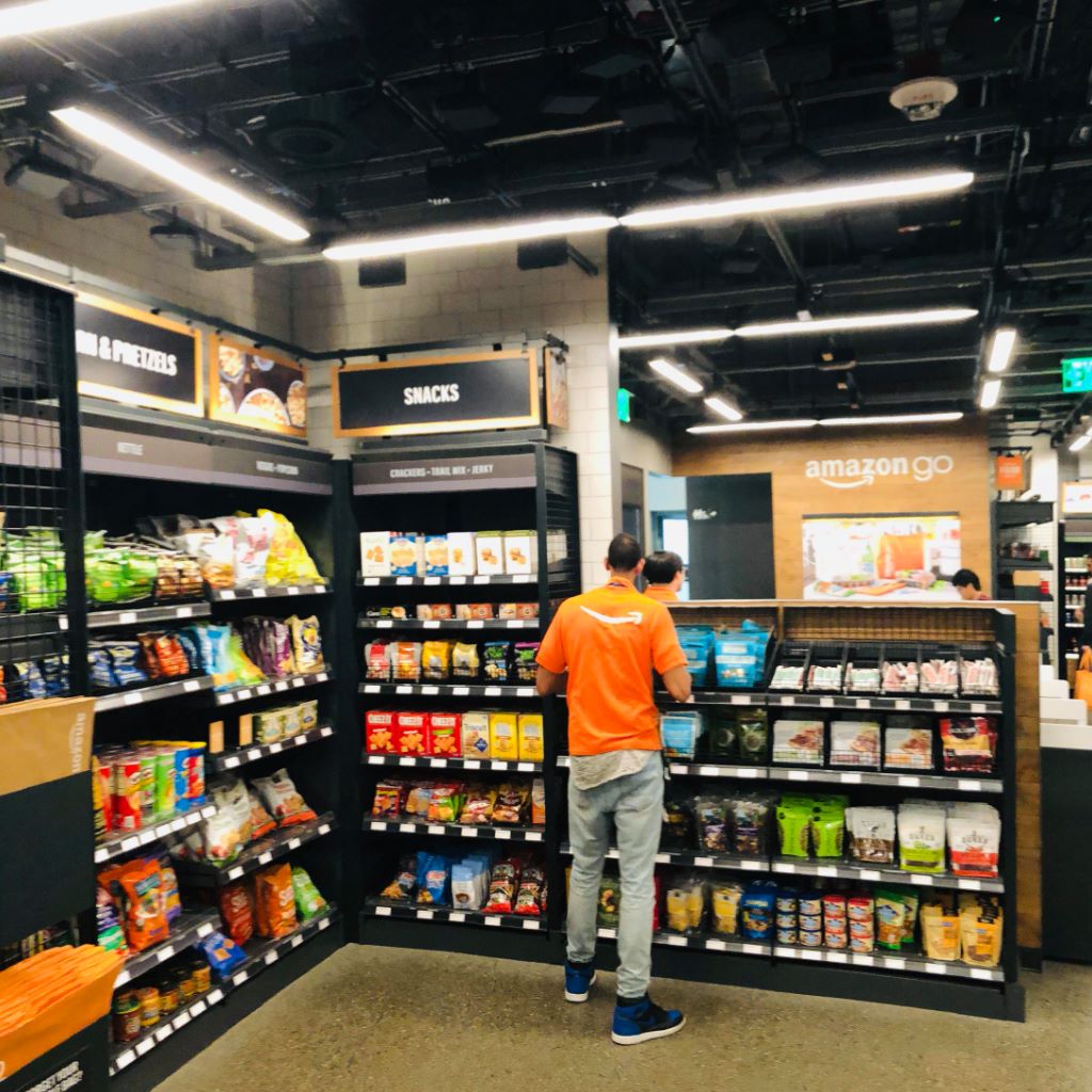 An Amazon Go employee restocking the snacks aisle, shelves stacked with chips, crackers, and trail mix.