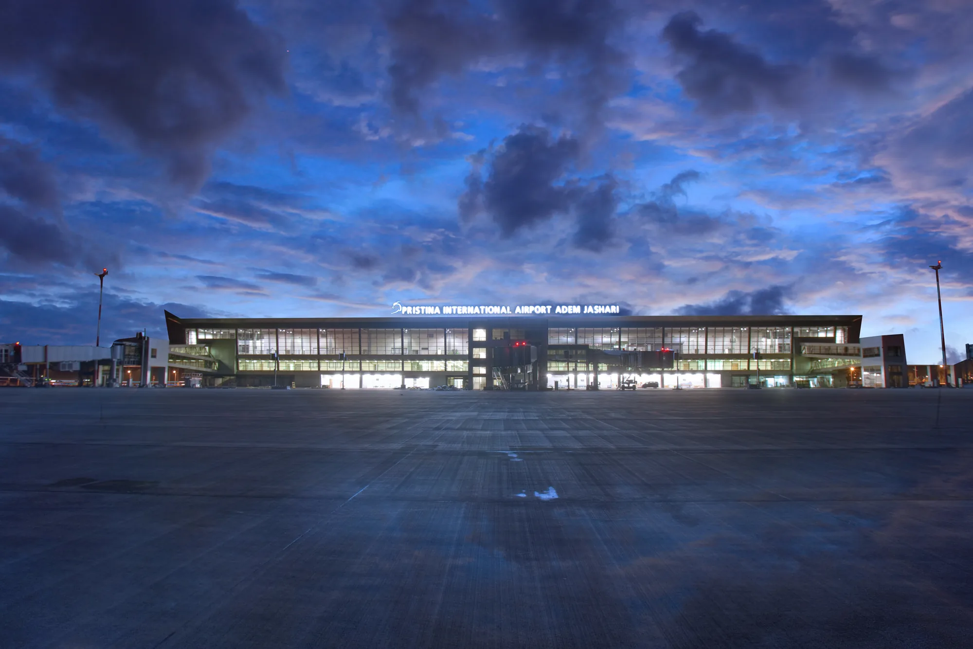 The arrivals hall at Prishtina International Airport Adem Jashari, with travelers walking past the terminal's glass facade.