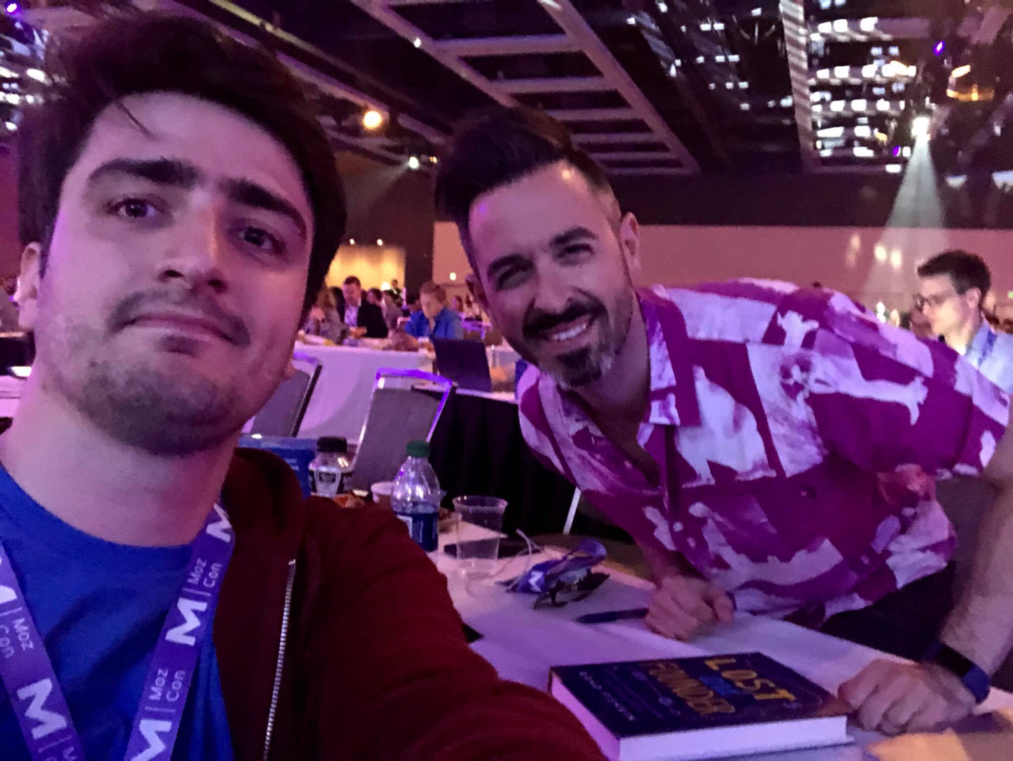 Me sitting next to Rand Fishkin at a table, his copy of Lost and Founder open in front of him as he signs it, at MozCon 2018.