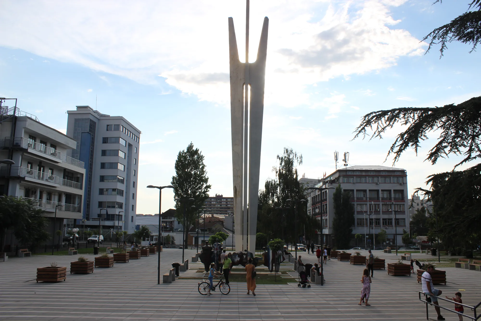 Adem Jashari Square in Prishtina, a wide paved plaza with people walking past surrounding buildings on a sunny day.
