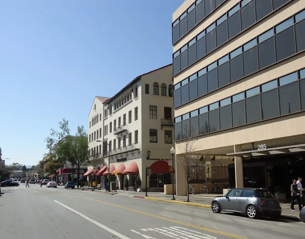 A quiet street in downtown Palo Alto lined with storefronts, parked cars and trees under a clear blue sky.
