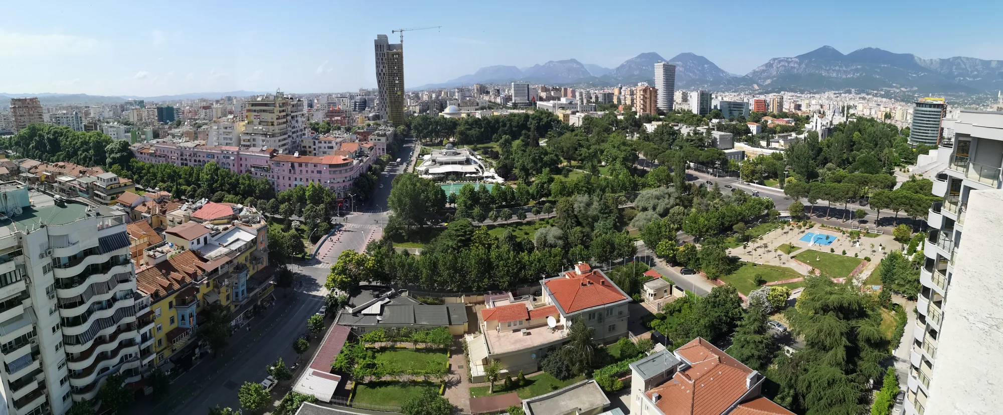 A panoramic view of Tirana, Albania, showing its dense mix of apartment blocks and green space under a wide sky.